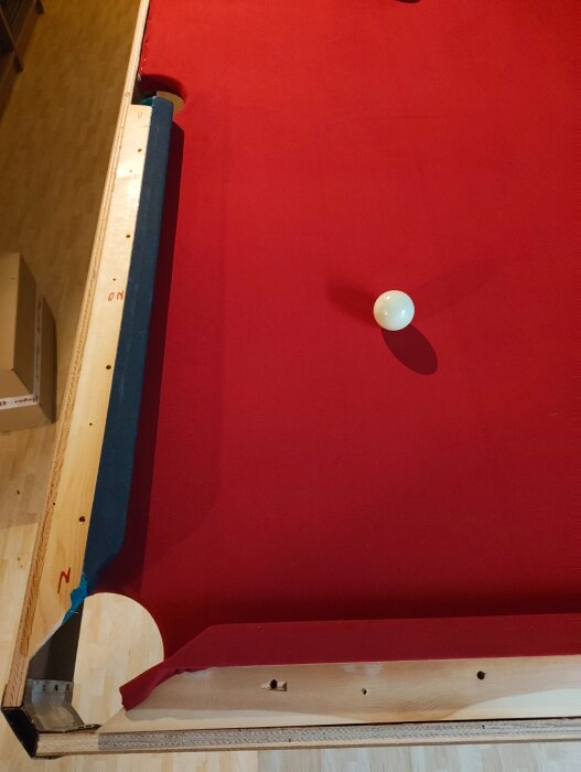 Biljardbord med röd filt och träkant under renovering, vit biljardboll i mitten och en ficka synlig längst ner.
