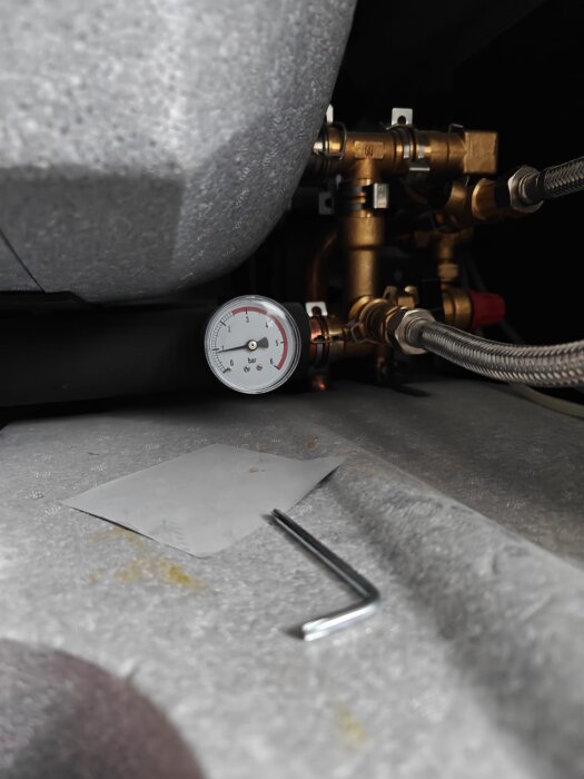 Manometer och röranslutningar på en värmepump, med en insexnyckel och papper liggande på golvet under.