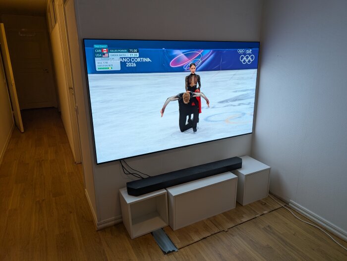 Väggmonterad TV visar konståkning, med vit TV-möbel och högtalare under, samt lösa kablar synliga på golvet.