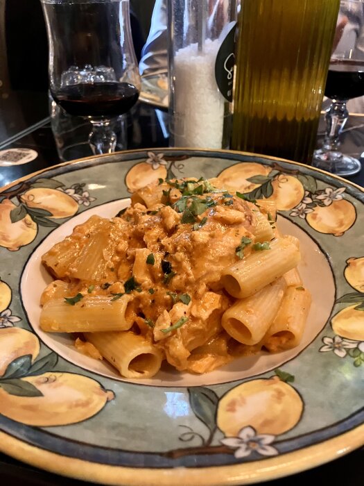 Pasta med lax serverad på en dekorerad tallrik, ackompanjerad av rött vin i ett glas i bakgrunden.