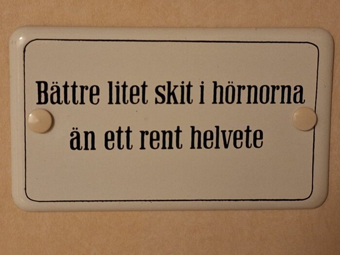Vit skylt med texten "Bättre litet skit i hörnorna än ett rent helvete" på beige bakgrund.