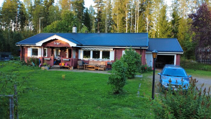 Rött hus och garage med blått plåttak, omgiven av grönska. En bil syns på uppfarten. Nyplatsat tak med Planja Regent-plåt över Shingeltak.