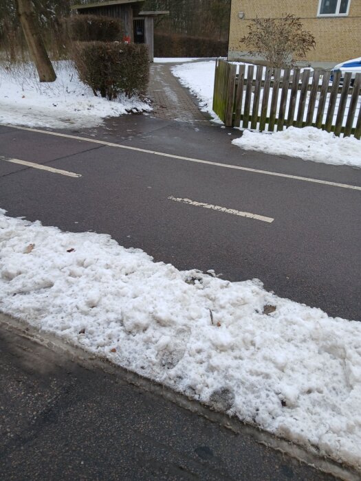 Snövall vid staket och trottoar som orsakar problem för renhållningstransport, skapad av plogbil, med snö täckande marken och vägen.