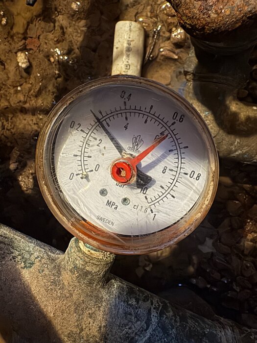Manometer med röda och svarta visare visar tryck i bar och MPa, fotograferad över rördelar, för diskussion om dåligt vattentryck hemma.