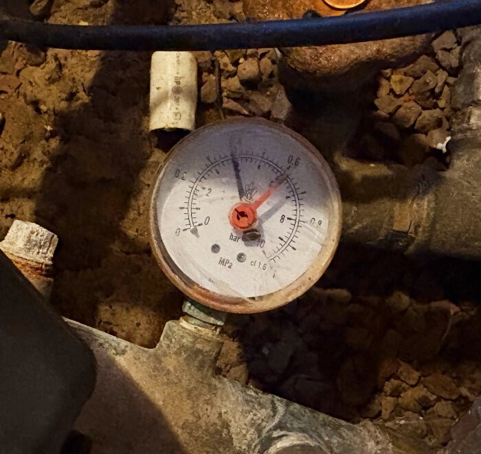 Manometer på rörsystem för vatten, med två nålar och streck för bar- och MPa-mätning, placerad på markig yta.