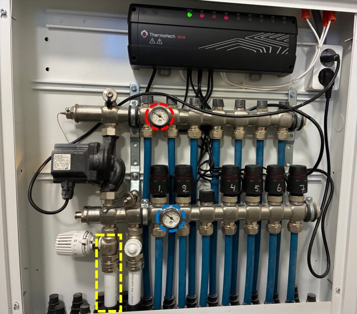 Värmepumpsinstallation med rör och temperaturmätare i ett skåp, röd och blå mätare visar olika grader, elektronisk styrbox upptill.