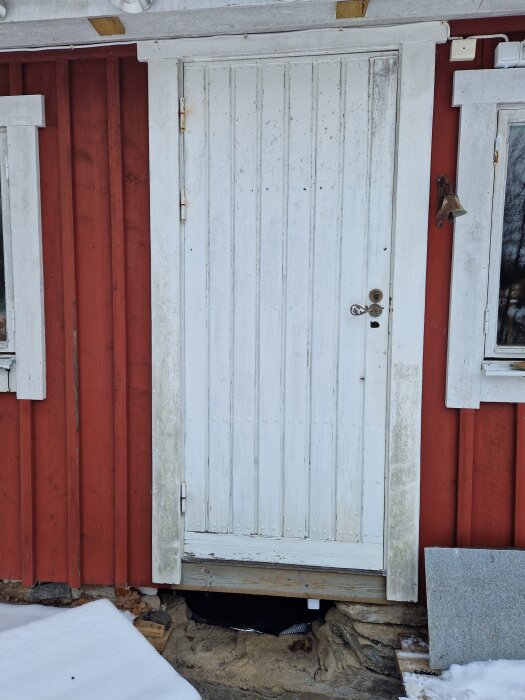 Sliten vit ytterdörr med äldre design monterad på röd panelvägg, omgiven av snö och sliten omgivning.