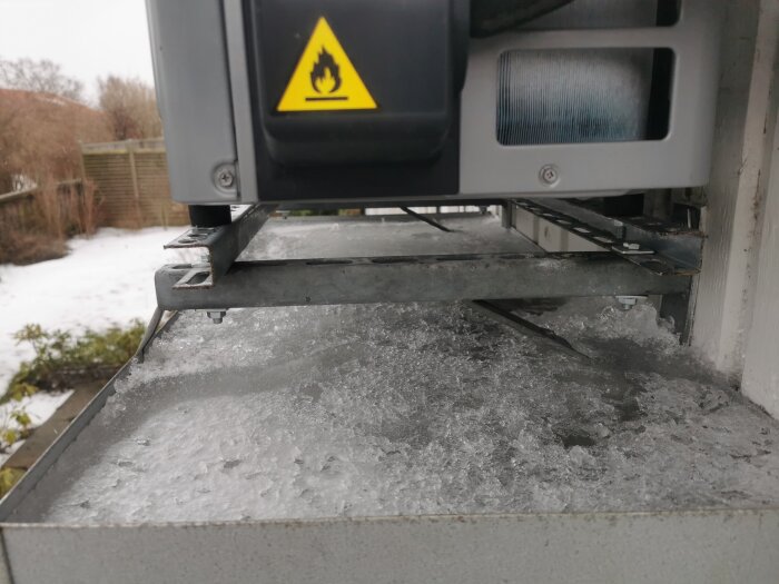 Utomhusanordning med isbildning och ett varningsmärke för brandrisk. Snötäckta buskar och staket syns i bakgrunden.