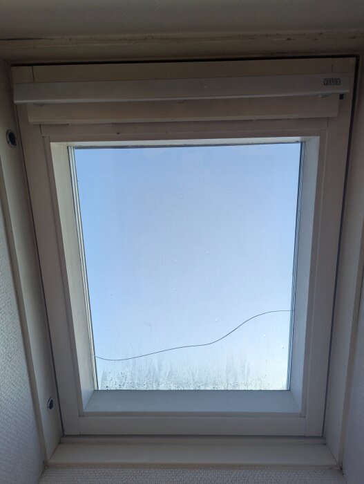 Velux takfönster med en horisontell spricka i glaset, sett från insidan mot en blå himmel.