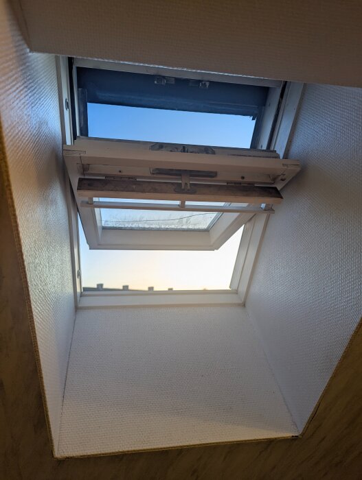 Öppet Velux takfönster i ett badrum, med träkarm och en horisontell spricka i glaset, insläppande blå himmel.
