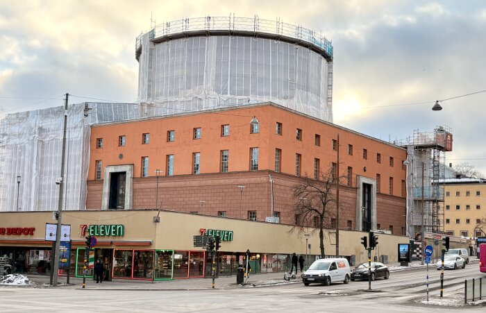 Stadsbiblioteket i Stockholm täcks av efter omputsning i brunrosaorange nyans, med byggställningar och närliggande 7-Eleven och trafik.