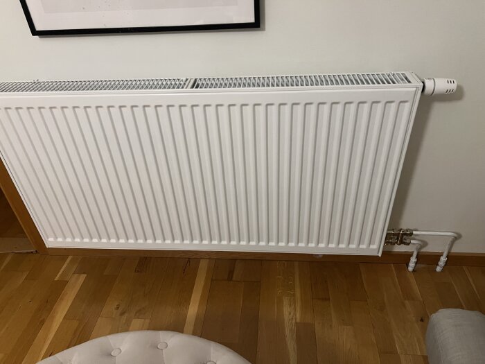 Vitt element monterat på en vit vägg, omgivet av träparkettgolv och en del av en beige möbel syns i nederkant av bilden.