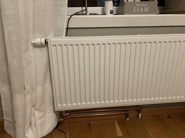 Vitt element under ett fönster, anslutet med kopparrör, omgivet av vita gardiner och träparkettgolv.