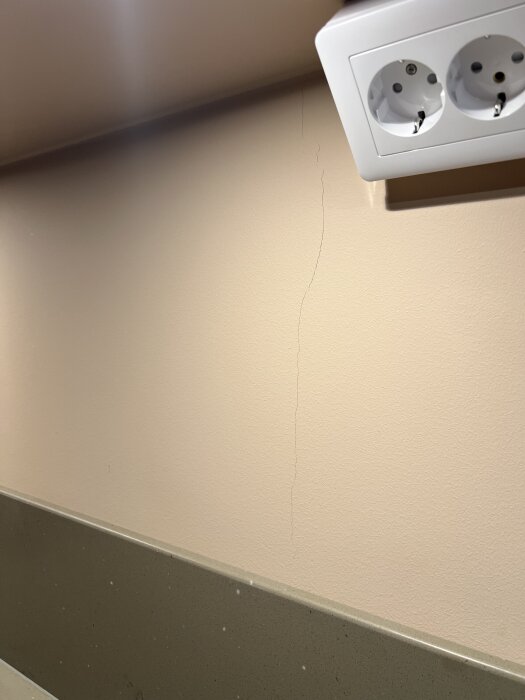 Spricka i nymålad köksvägg nära ett dubbelt vägguttag, ovanför en stänkskydd vid köksbänken.
