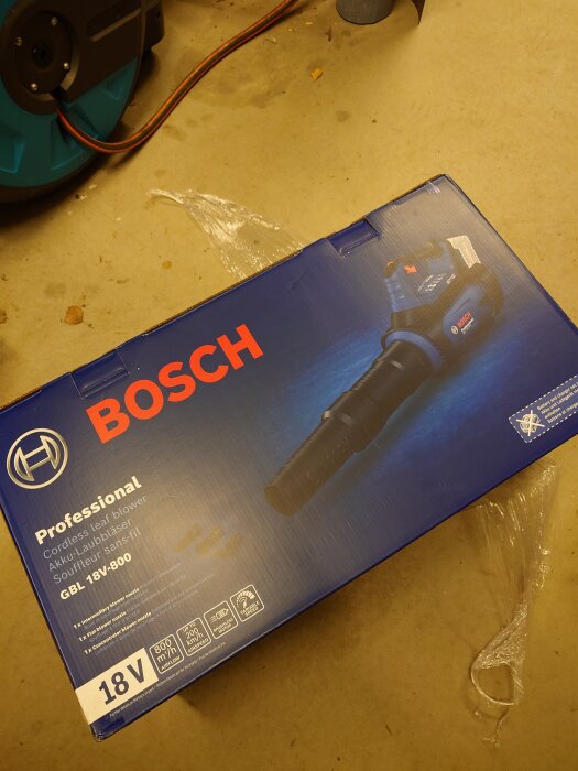 Kartong med Bosch cordless lövblåsare, modell GBL 18V-800, på en golvyta i ett garage eller verkstad.