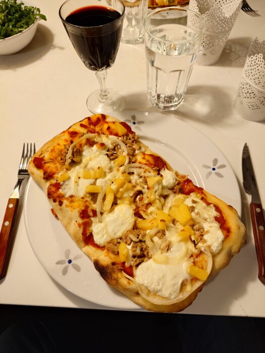 Pinsa med toppings av tonfisk, burrata, lök, ananas och mozzarella serverad med ett glas rött vin och ett glas vatten.