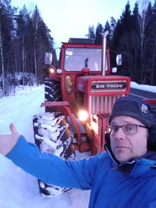 Man i blå jacka poserar framför en röd BM Volvo traktor på en snötäckt väg, omgiven av skog.