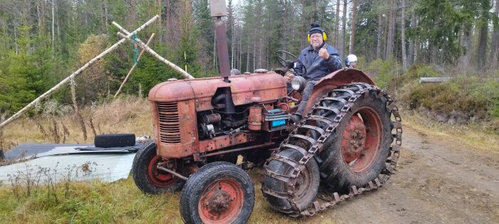 En man sitter på en röd Volvo T24 traktor från 1952 på en skogsväg, omgiven av träd och kvistar.