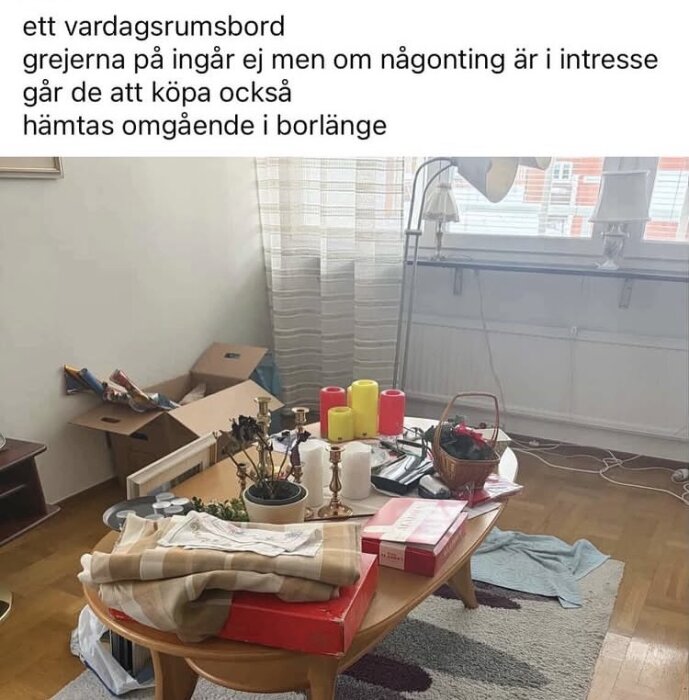 Ett vardagsrumsbord med diverse föremål, inklusive en Aladdinask, ljus, textilier och växter, mot en fönsterbakgrund.