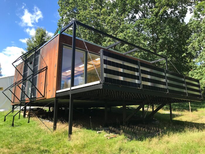 Modern byggnad med stora glaspartier och svart konstruktion på upphöjda metallben, omgiven av grönska och träd.