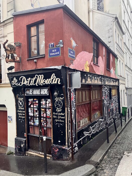 Röd byggnad på Rue Tholozé, dekorerad med graffiti och skyltar, med texten "Le Petit Moulin" och ett antal klistermärken på dörren.