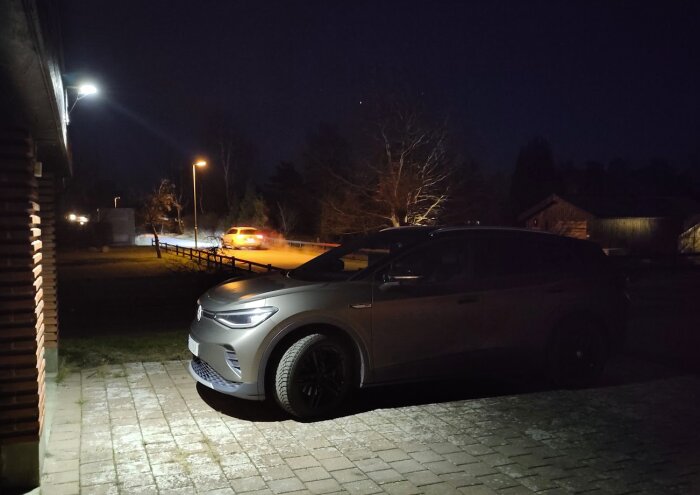 Bil parkerad på uppfart på kvällen med gatubelysning och ett annat fordon synligt i bakgrunden.