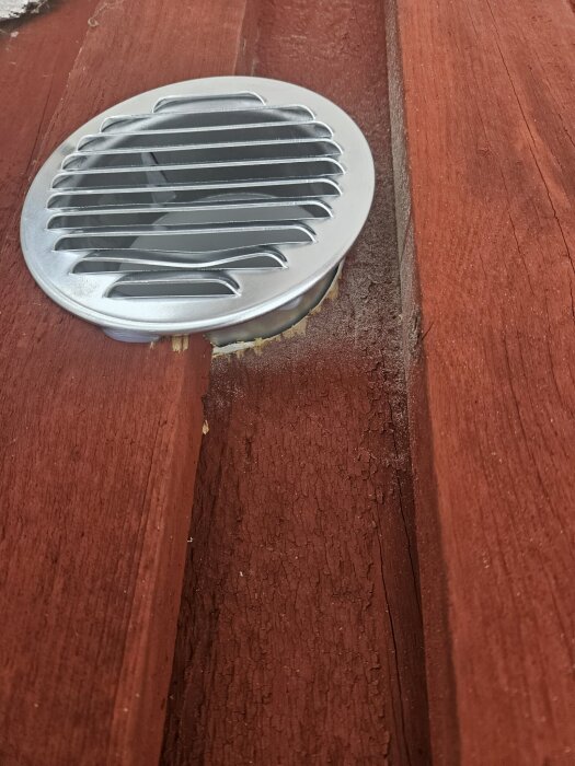 Silverfärgat ventilgaller monterat utanför den rödmålade panelen, med synligt mellanrum mot brädan.