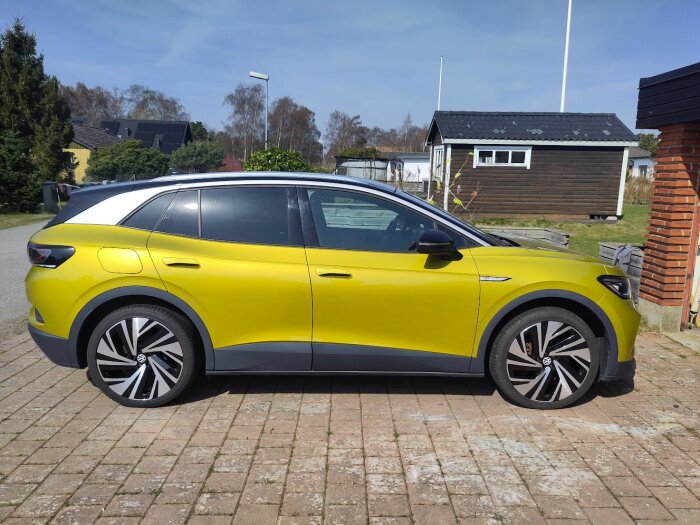 Gul bil parkerad på en uppfart framför ett hus, med träd och en garagebyggnad i bakgrunden.