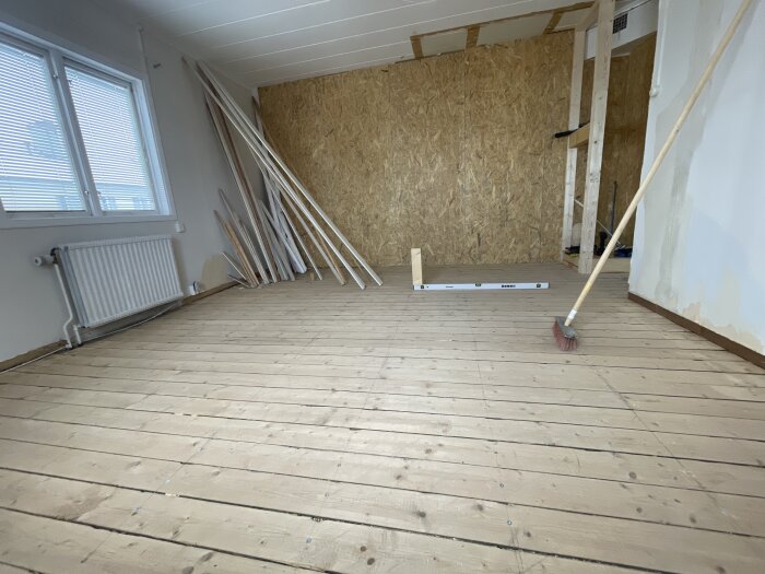 Rum med spontade golvbrädor, ett fönster, en radiator och en stapel brädor lutad mot en vägg. Ett vattenpass ligger på golvet.