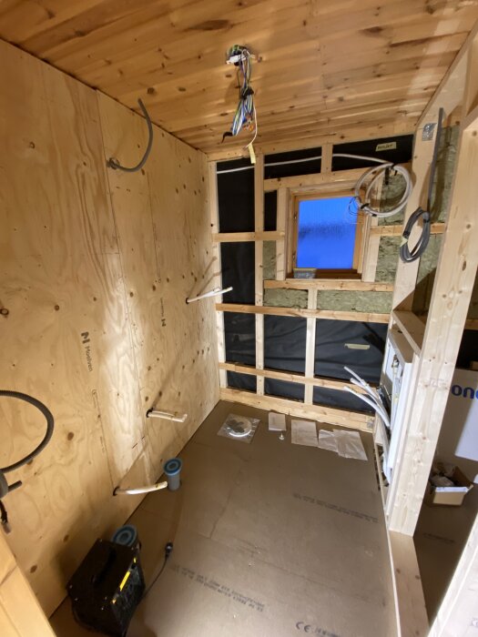 Oisolerat rum med träpanel, upphängda kablar och rördragning; byggprojekt pågår. Fönster på väggen. Verktyg och byggmaterial placerade på golvet.