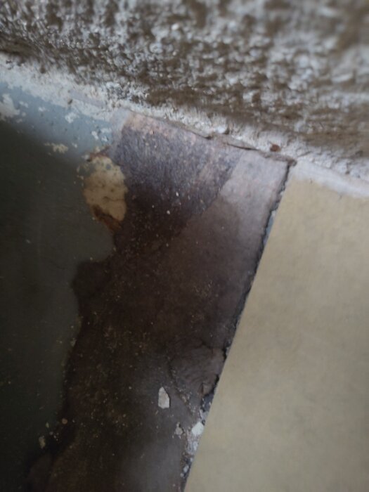 Närbild av golvskikt i källare, med en mörk filtliknande matta mellan linoleum och betong. Fråga om asbestinnehåll.