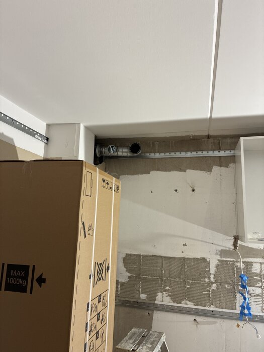 Vägg med en metallventil i ett rum under renovering, kartong märkt "MAX 1000kg" står intill väggen, byggmaterial synligt.