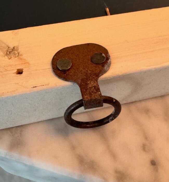 Rostigt upphängningsbeslag monterat på träbit, med metallring och två nitar, placerat på marmorunderlag.