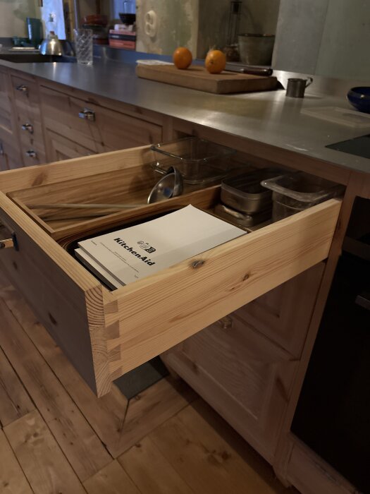 Öppen kökslåda i trä med handbok och köksredskap. Lådan är del av en köksinredning i trä med rostfri bänkskiva.