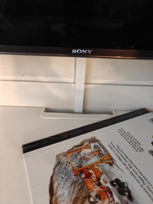 TV med Sony-logga monterad på ljus träpanel med kabelkanal, bok med illustration ligger på bordet nedanför.