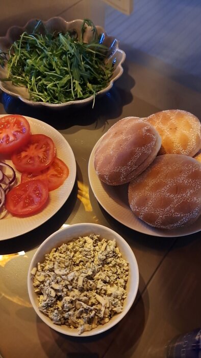 Hamburgeringredienser: skålar med ruccola, tärnad gräddädelost, samt fat med skivade tomater, rödlök och hamburgerbröd.