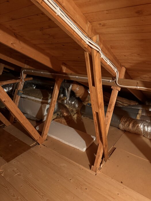 Vindsutrymme i ett hus med träbalkar, isolering och ventilationsrör synliga.