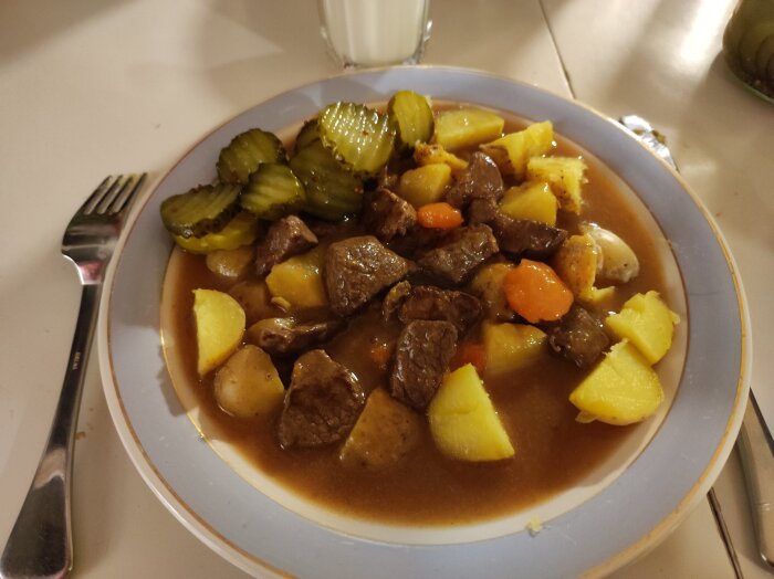 Tallrik med köttgryta, potatis, morötter och skivade inlagda gurkor, serverad med en dryck i bakgrunden.