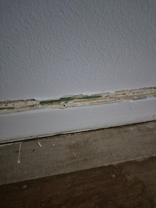 Närbild av en vägg där tapet och färg lager avlägsnats nära golvlisten, avslöjande gamla lager i olika färger och texturer.