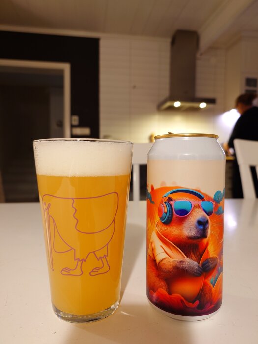 Ett glas med ljus öl och en burk med färgstarkt motiv av ett djur med hörlurar och solglasögon, på ett köksbord.