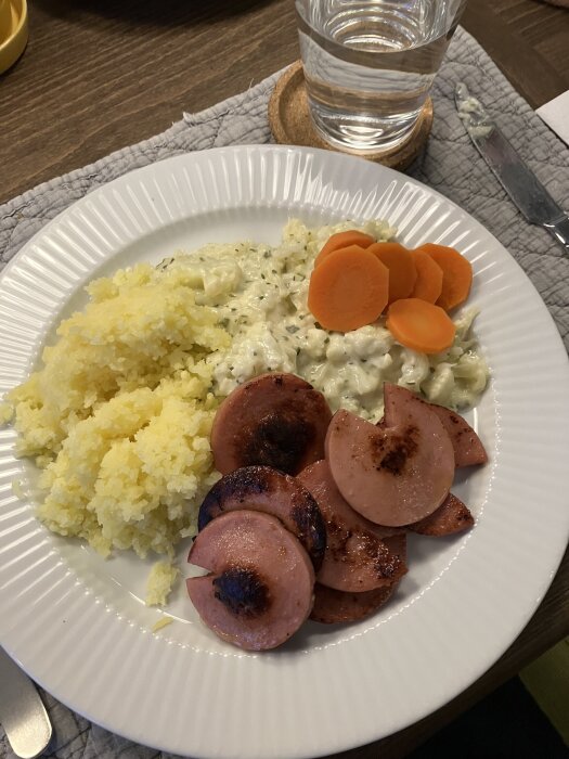 Tallrik med stekt korv, potatismos, stuvade grönsaker och kokta morötter. Ett glas vatten finns bredvid på en bordstablett.