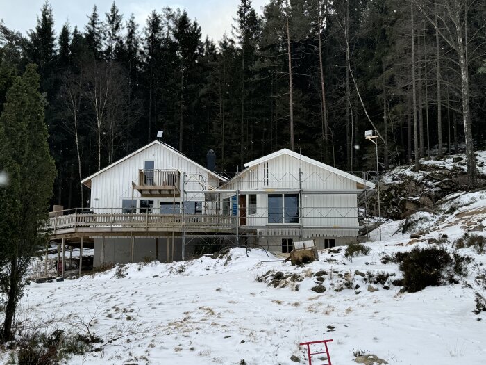 Nybyggt hus med altan och skjutdörrar, omgivet av snötäckt mark och skog i bakgrunden. Scenen visar vintrig miljö vid ett byggprojekt.