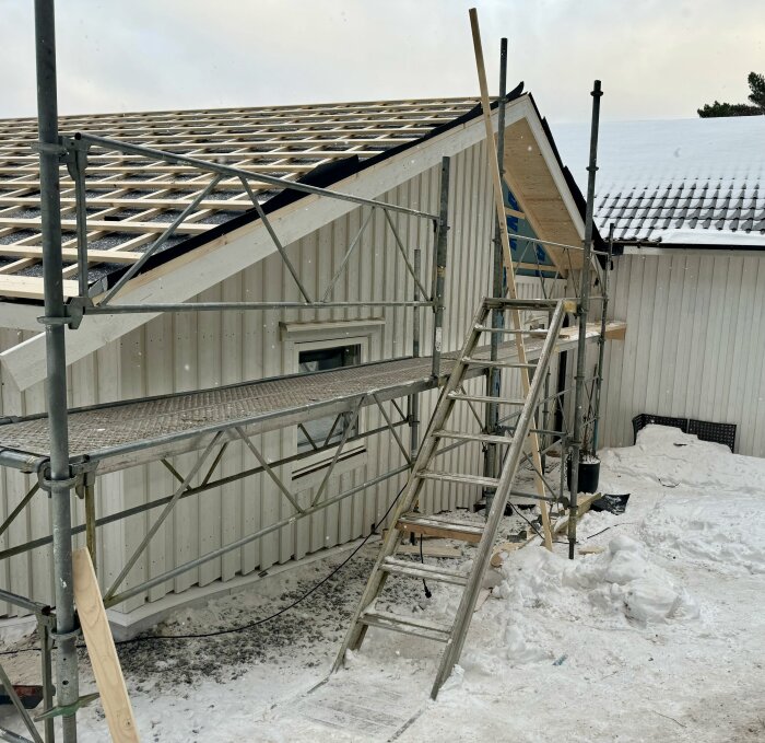 Byggarbetsplats för hus under vinterväder, med byggnadsställning och stege vid husets tak, snö på marken.