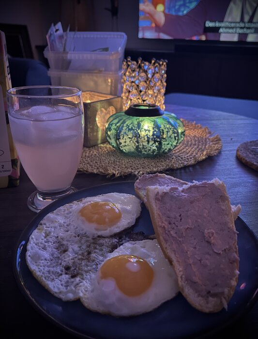Tallrik med två stekta ägg och bröd med leverpastej bredvid ett glas isvatten. Bakgrund med levande ljus och tända lampor.