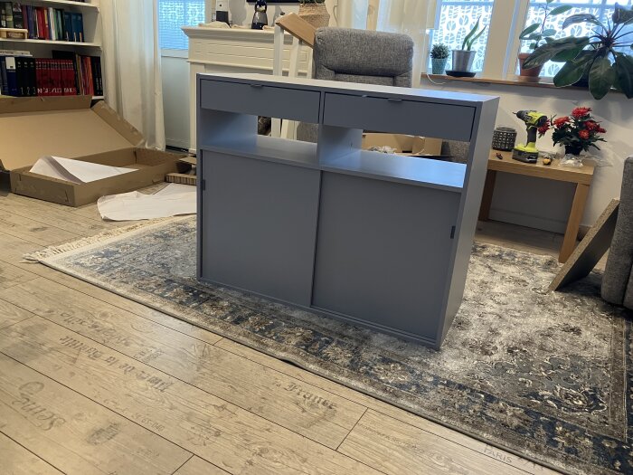 Grå IKEA-skåp på trägolv med öppningar på golvsidan och arbetsyta ovanpå, omgiven av verktyg och inbyggnadsdelar, hemma i ett vardagsrum.