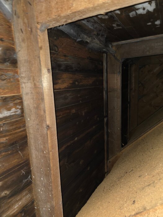Kallvind med träbjälkar och smutsig isolering, antydan till mikrobiell tillväxt på ytorna, mörkt och gammalt trä från ett hus byggt på 1940-talet.