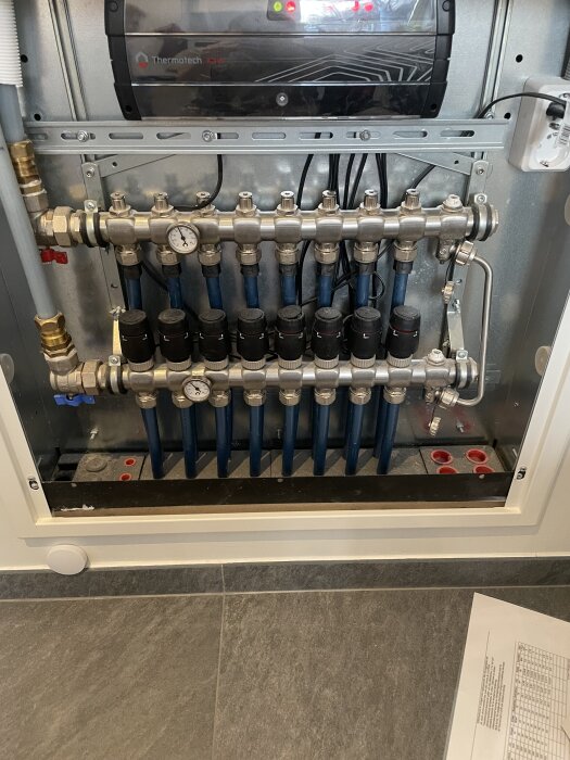 Värmefördelare i ett skåp med flera rör och temperaturmätare, del av ett radiator- eller golvvärmesystem.