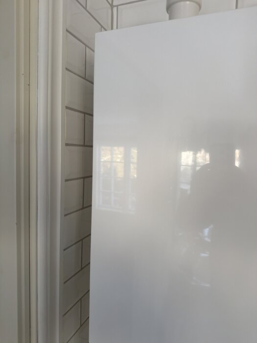 Glänsande vit yta av apparat eller panel i ett rum med kakelvägg. Reflektion av ett fönster syns på ytan.