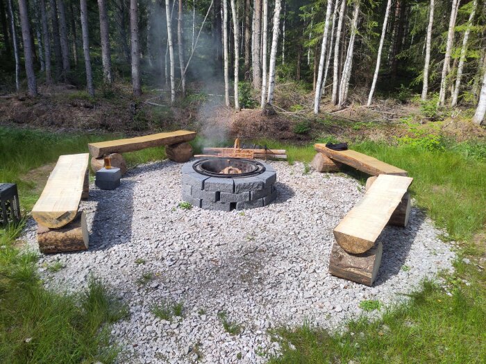 En grillplats med en ring av kantsten, sittbänkar av stockar och grusunderlag, omgiven av träd och grönska.