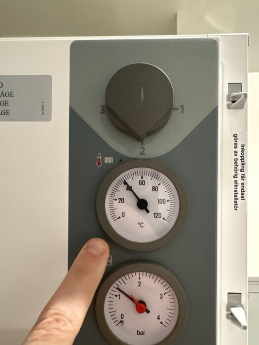 Termostat och två mätare på en Nibe EVC13, med hand som pekar på temperaturmätare som visar cirka 58 grader Celsius.
